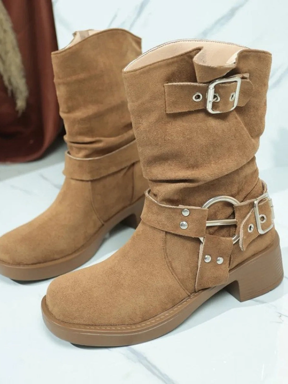 Tan suede-look boots feature round toes, low block heels, slouchy shafts, wraparound straps, and silver buckle hardware for a rugged style.