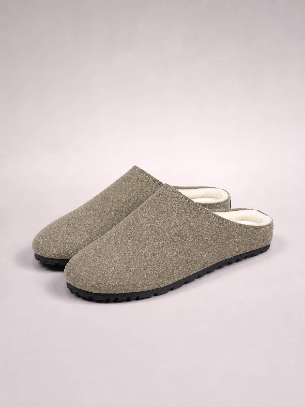 Taupe suede round-toe flat slipper mules feature a backless design, plush lining, stitched edges, and sturdy black rubber soles.
