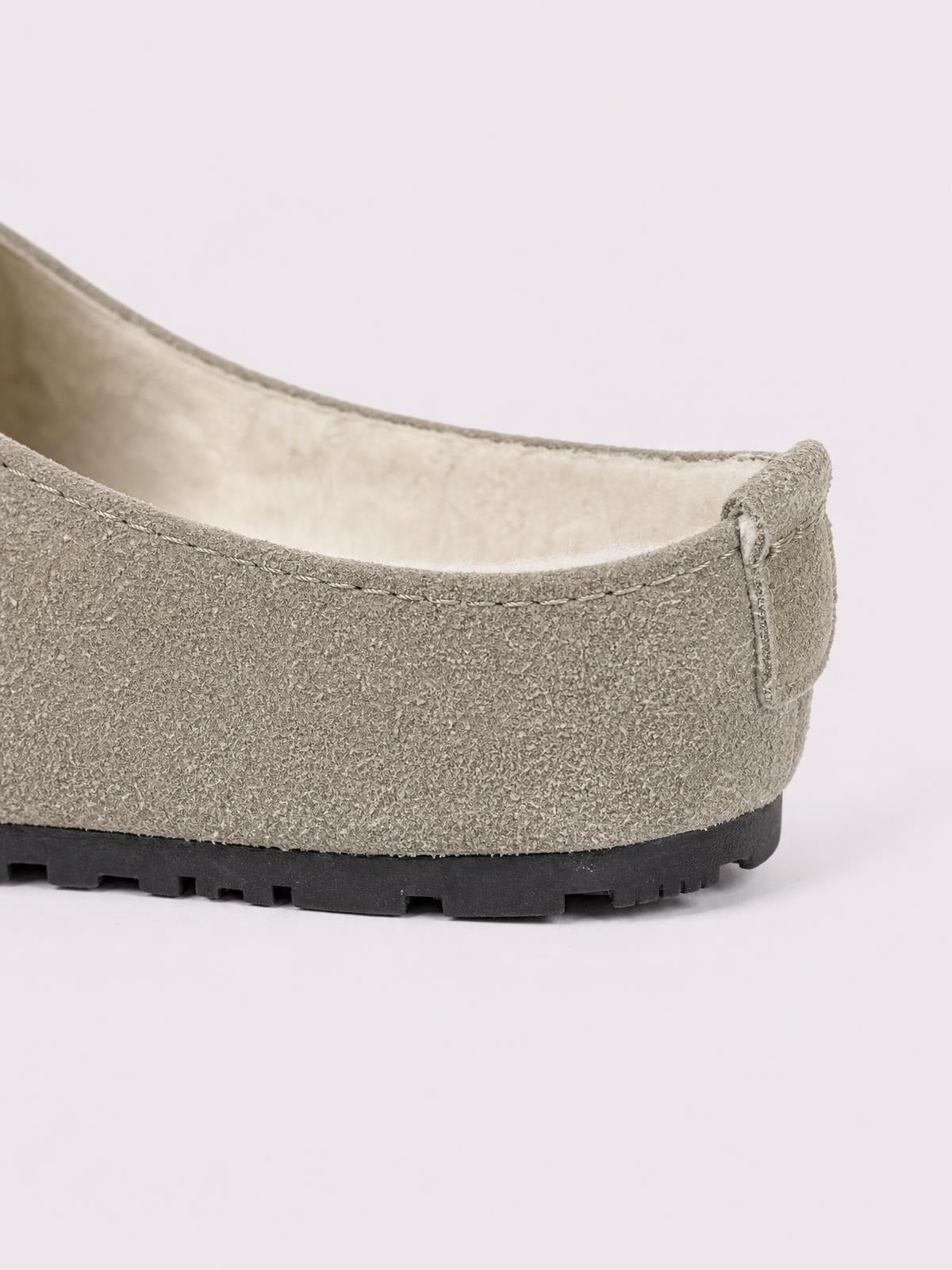 A taupe suede flat slipper features a softly rounded silhouette, visible stitched construction, a plush light lining, and a black rubber lug sole.
