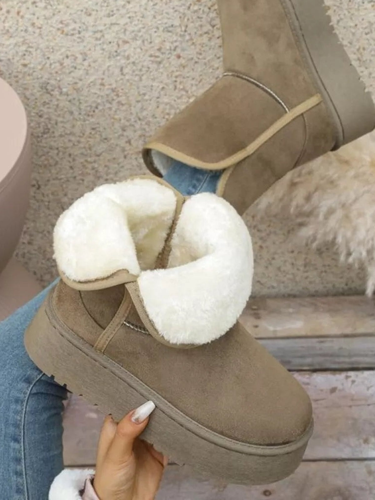 A hand holds taupe round toe platform boots with a thick flat rubber sole, ankle-height silhouette, and soft white fluffy lining over blue jeans.