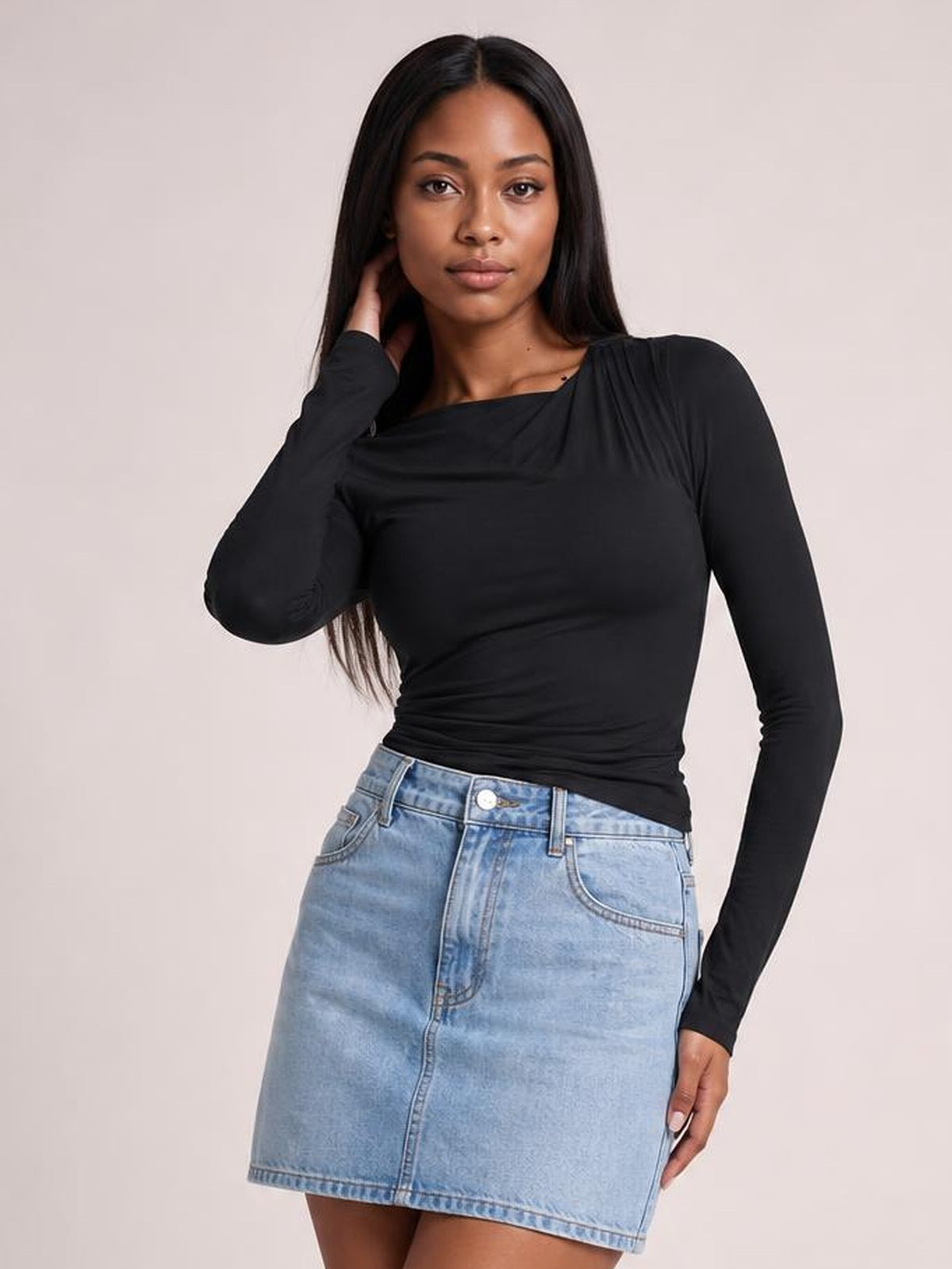Model poses with one hand at her hair, wearing a black ruched asymmetrical-neck long-sleeve T-shirt with a fitted silhouette and light-wash denim skirt.