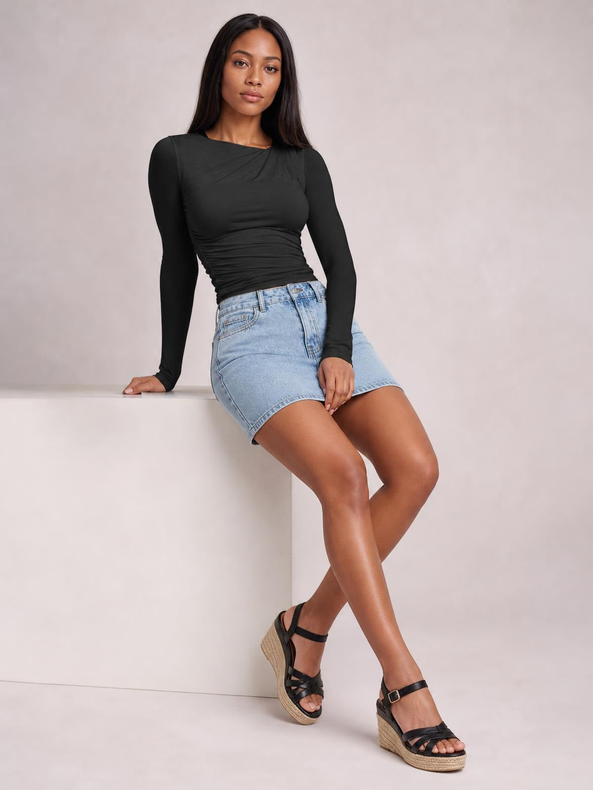 A model sits in a fitted black long sleeve T-shirt with an asymmetrical neckline, gathered ruching across the bodice, and a sleek stretch silhouette.