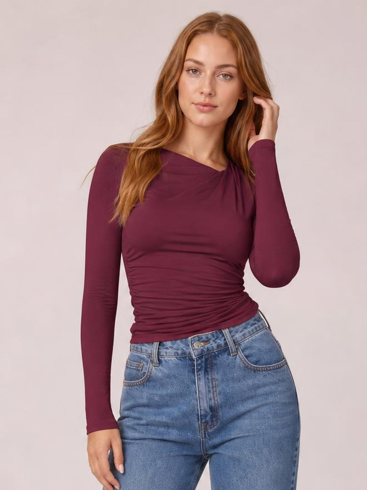 Model wears a burgundy ruched asymmetrical neck long sleeve T-shirt with a fitted silhouette, gathered waist, and one hand touching her hair.