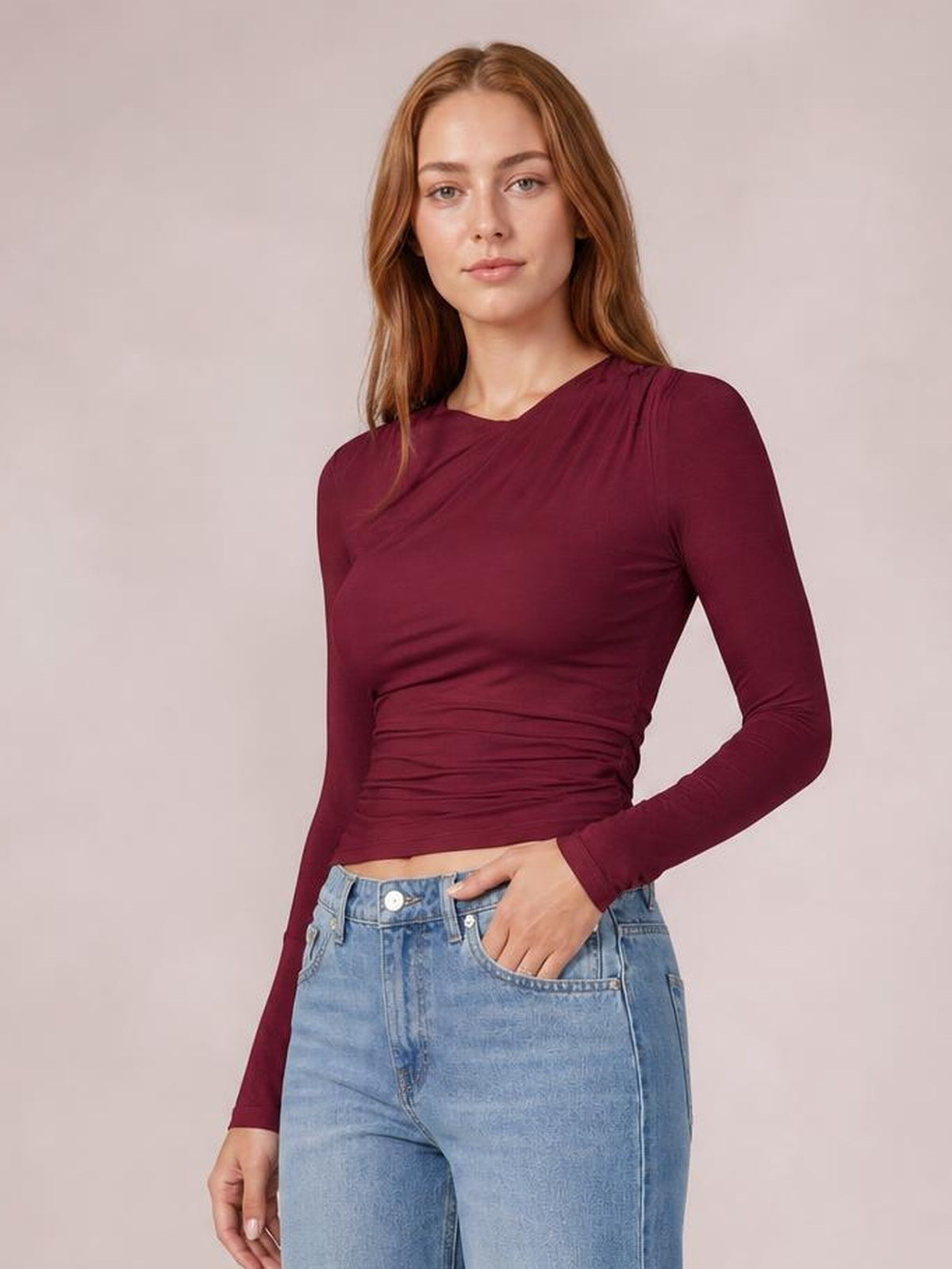 Model wearing a burgundy ruched asymmetrical neck long sleeve T-shirt with a fitted silhouette and gathered waist, one hand in her jeans pocket.