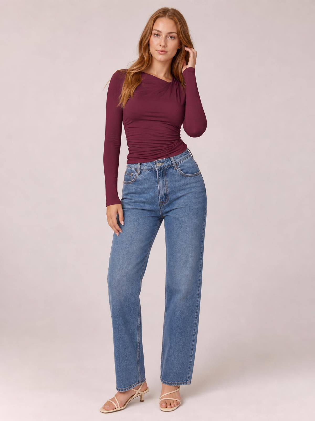 Model poses in a burgundy ruched asymmetrical-neck long-sleeve T-shirt with a fitted silhouette and gathered waist.