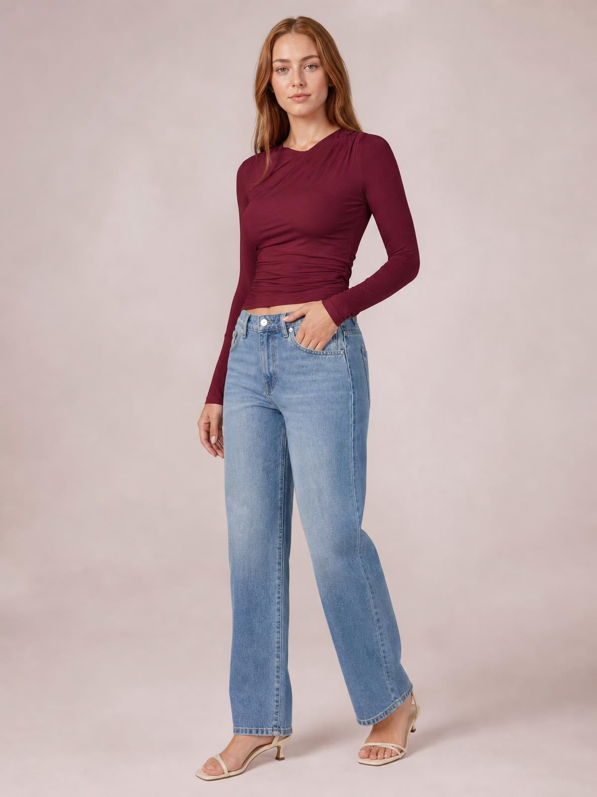 Model wearing a burgundy ruched asymmetrical neck long sleeve T-shirt with a fitted silhouette, posed with one hand in her jeans pocket.