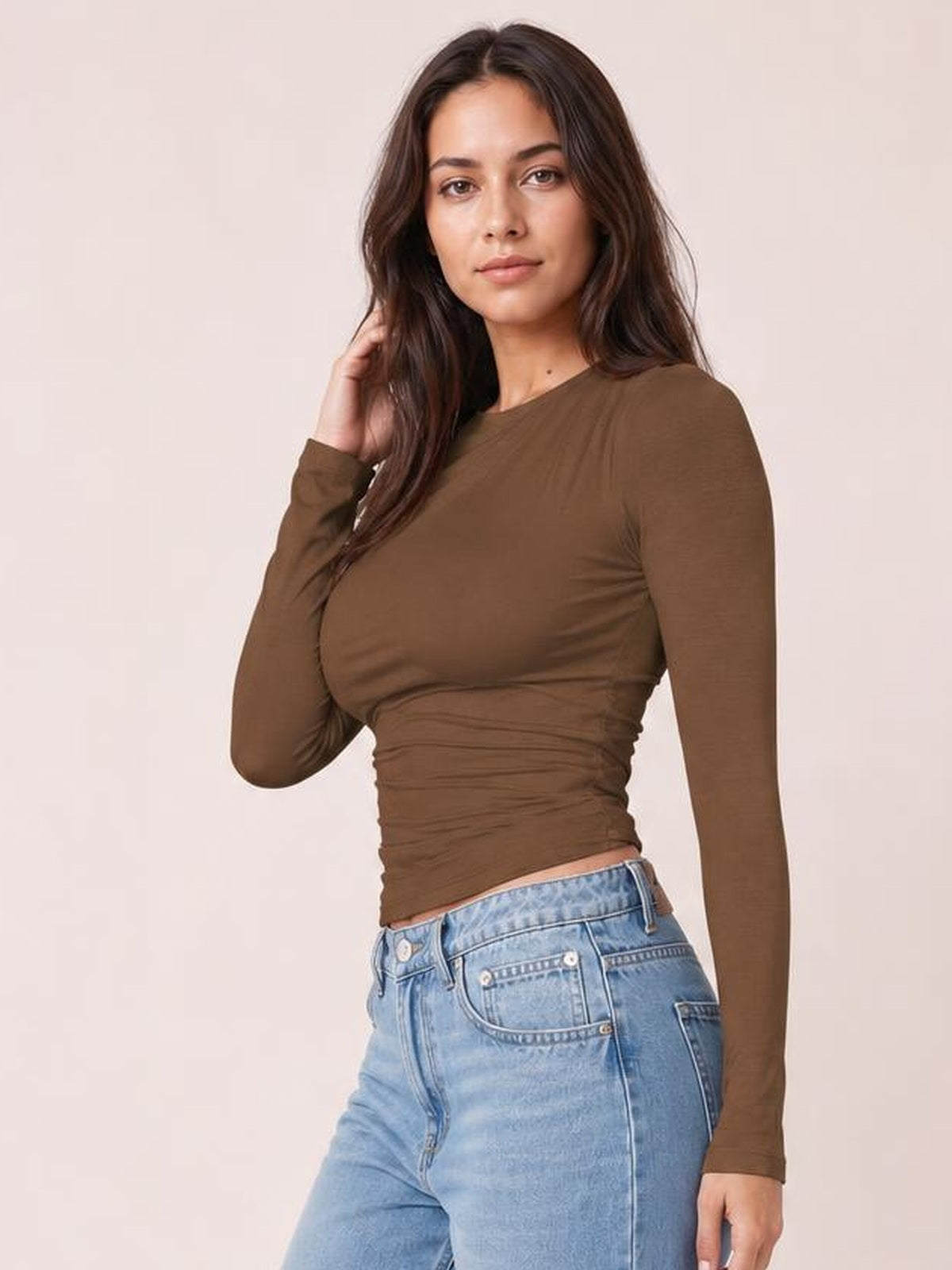A model poses in a brown ruched asymmetrical-neck long-sleeve T-shirt with a fitted silhouette and gathered detailing through the waist.