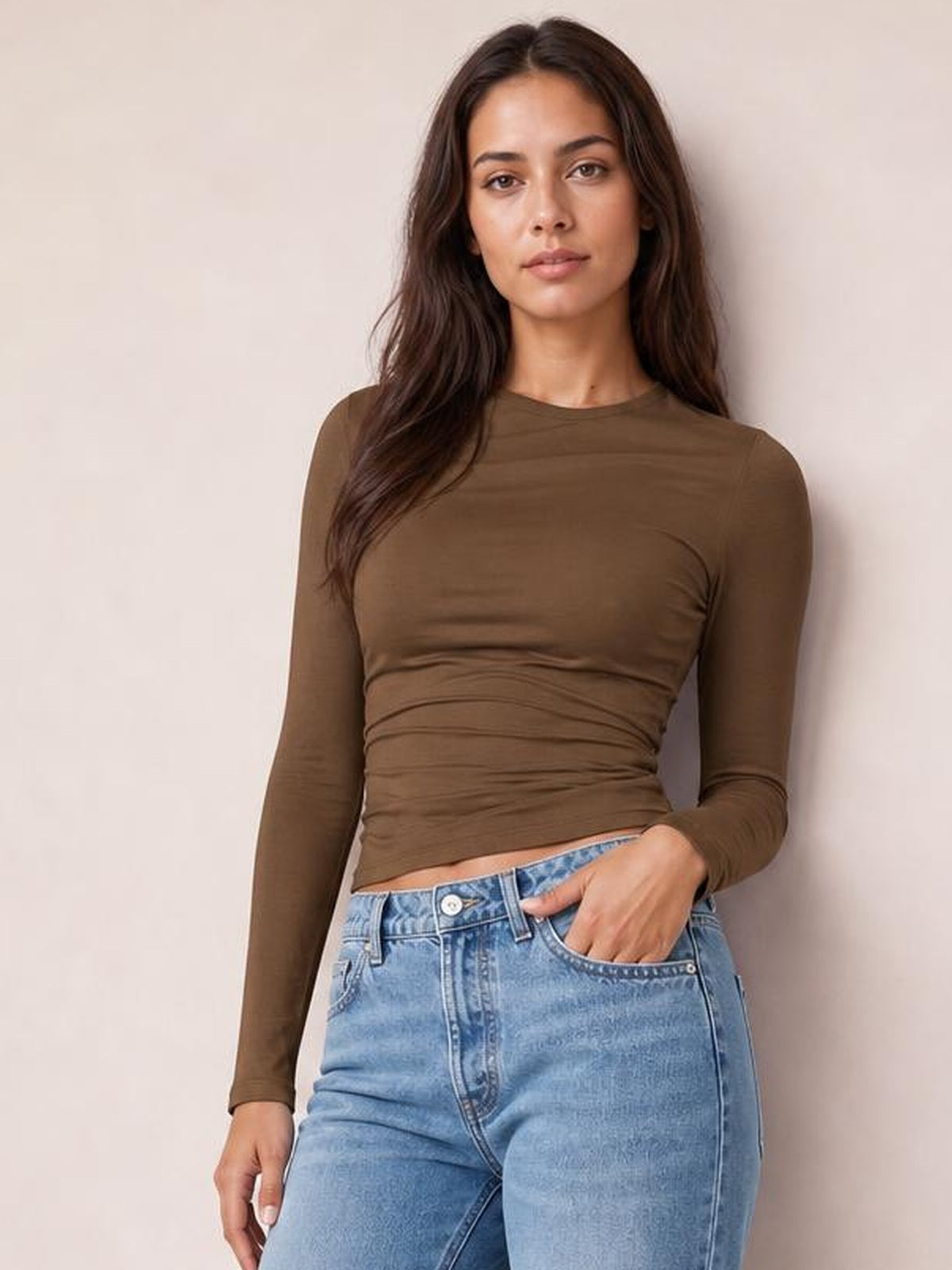 Model wears a fitted brown ruched asymmetrical-neck long-sleeve T-shirt with a smooth stretch silhouette, posing with one hand in her jeans pocket.