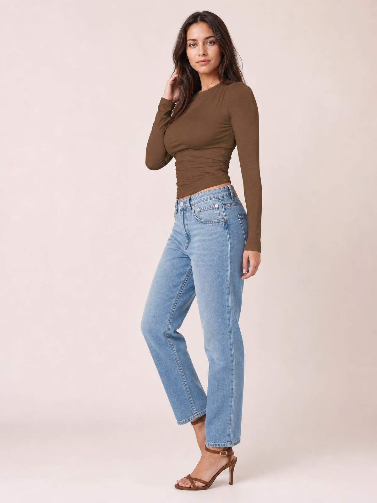 Model poses in a fitted brown ruched asymmetrical-neck long-sleeve T-shirt with a cropped silhouette and gathered side construction.