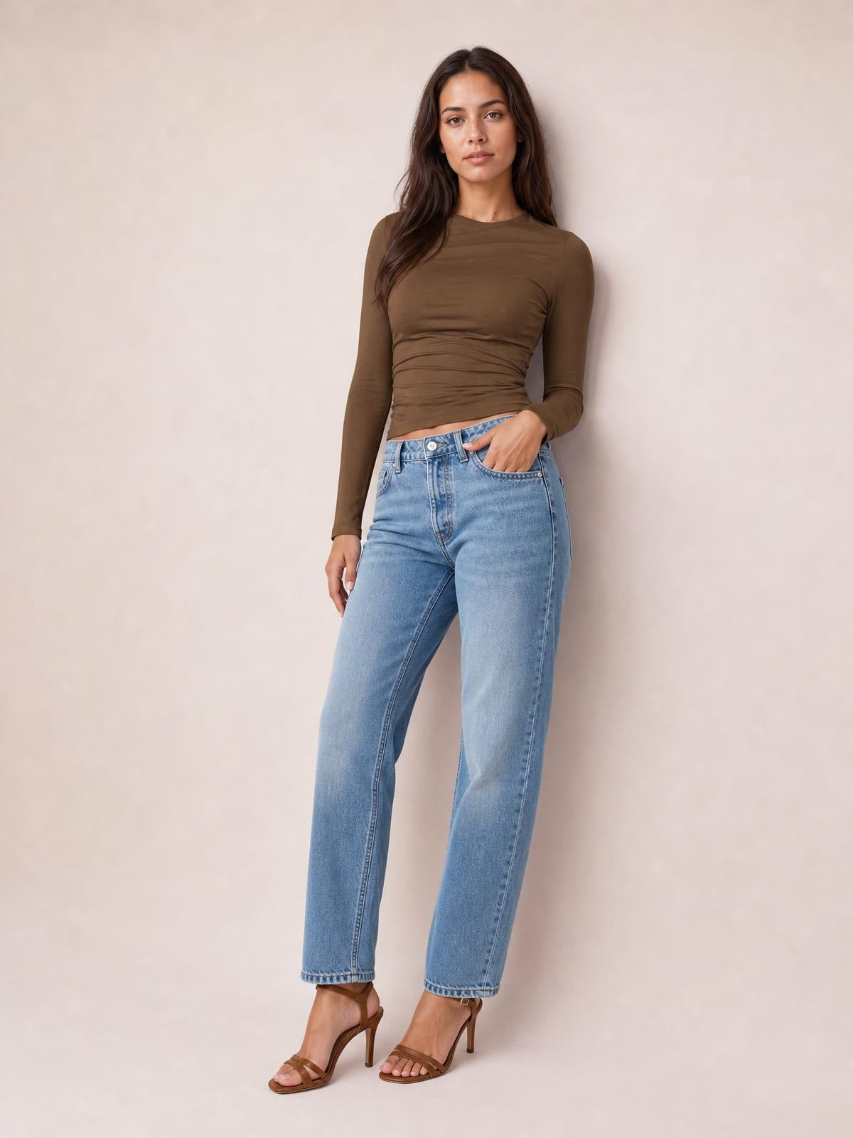 Model wears a brown ruched asymmetrical neck long sleeve T-shirt with a fitted, slightly cropped silhouette, posing with one hand in her jeans pocket.