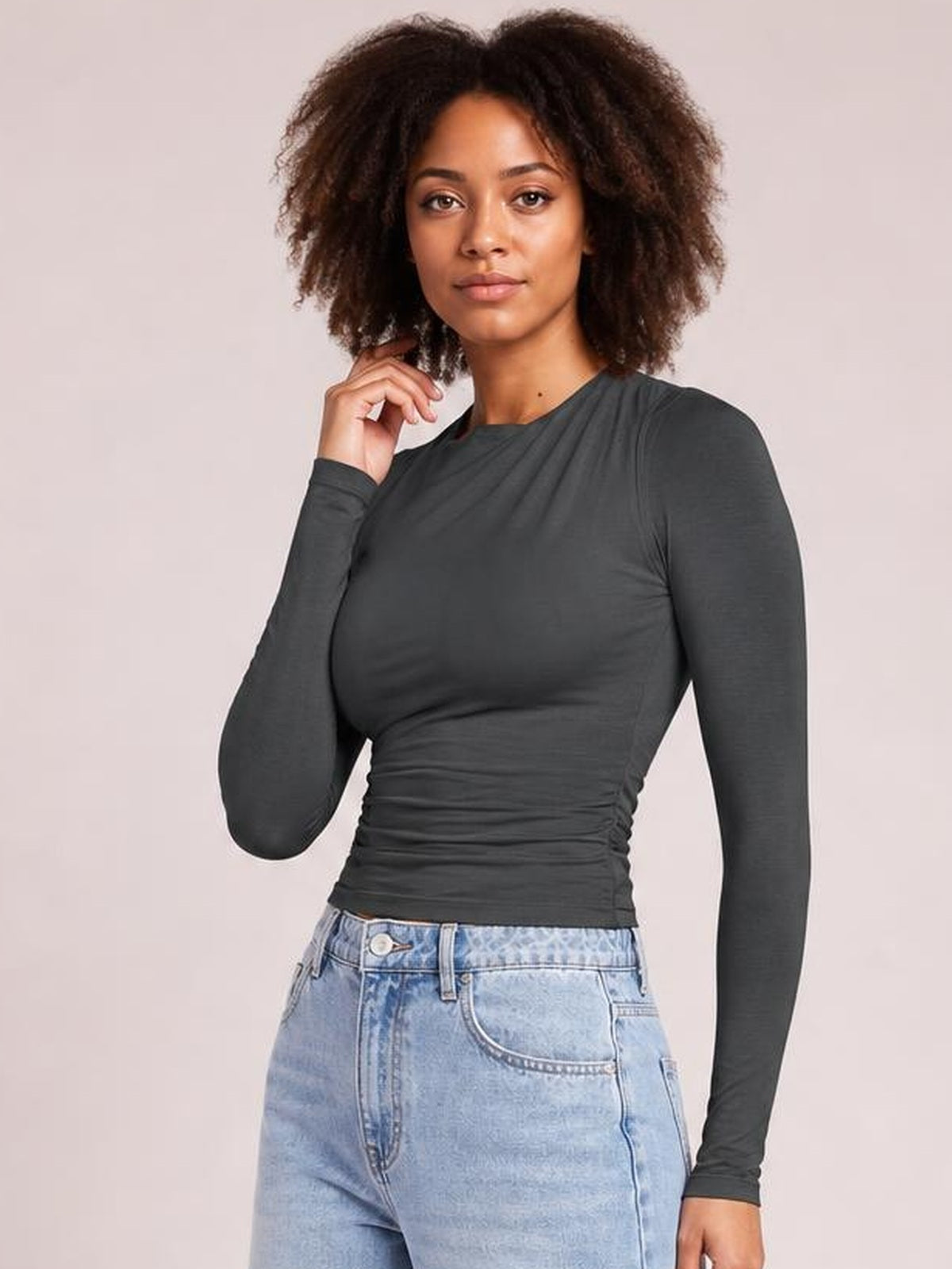 Model poses with one hand near her neck, wearing a charcoal ruched asymmetrical-neck long-sleeve T-shirt with a fitted silhouette and gathered waist.