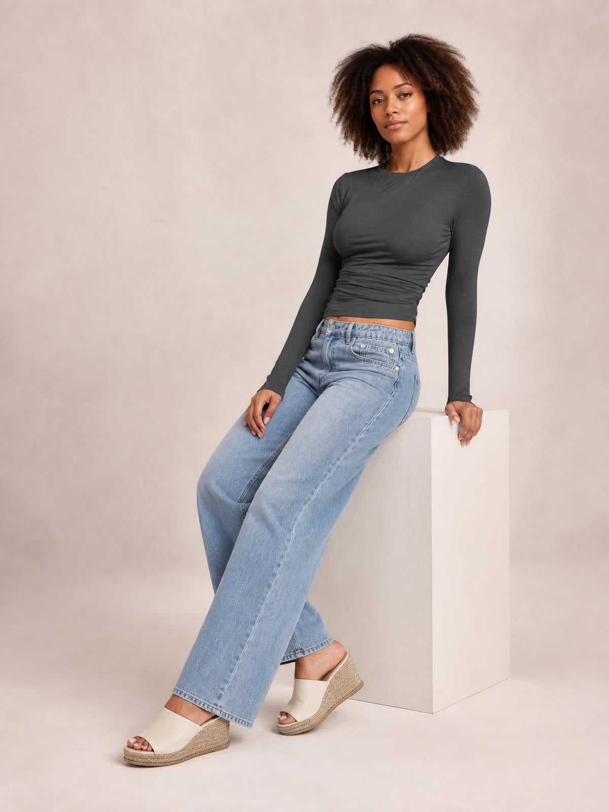 A model leans against a pedestal wearing a dark gray ruched asymmetrical-neck long-sleeve T-shirt with a fitted, cropped silhouette.
