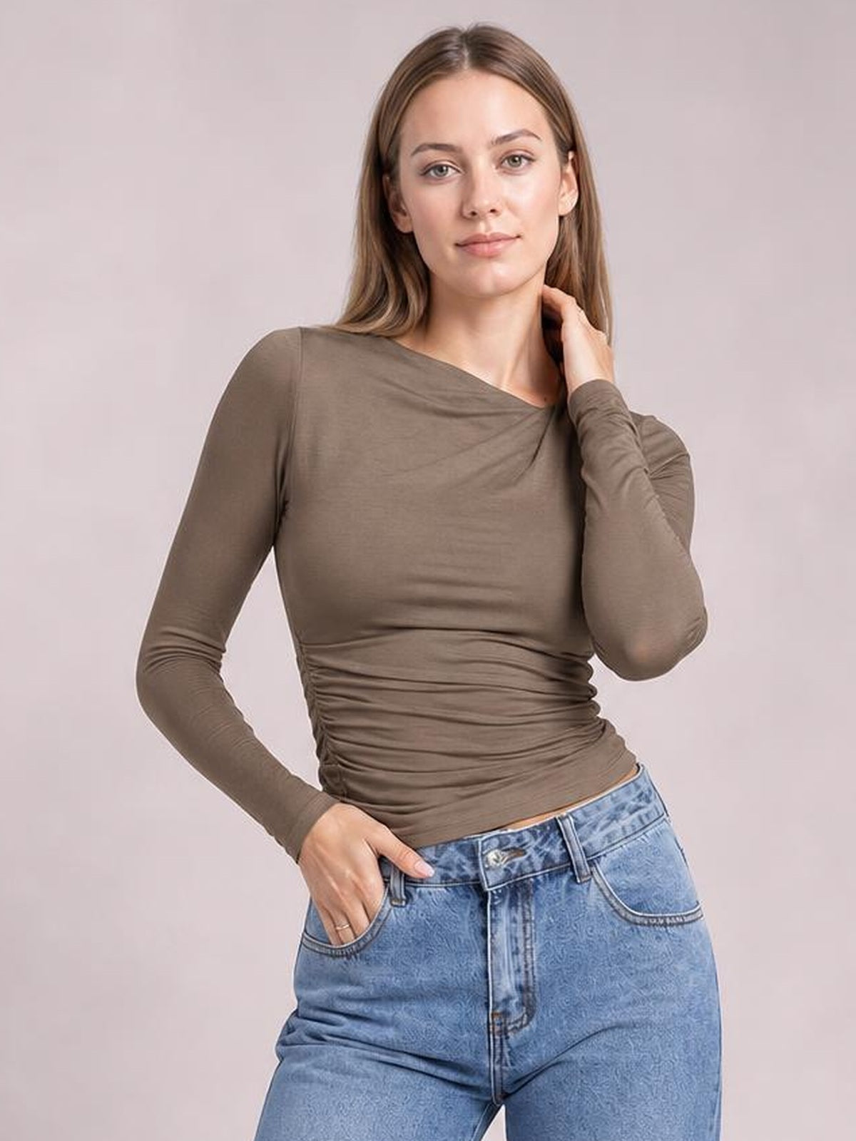 Model poses in a taupe ruched asymmetrical-neck long-sleeve T-shirt with a fitted silhouette and gathered construction through the waist.