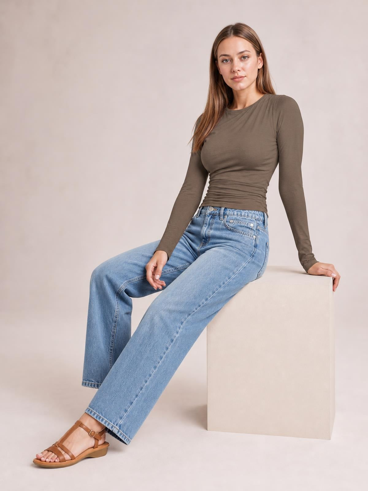 A seated model wears a taupe long-sleeve T-shirt with a fitted silhouette, ruched sides, and a softly gathered waist.