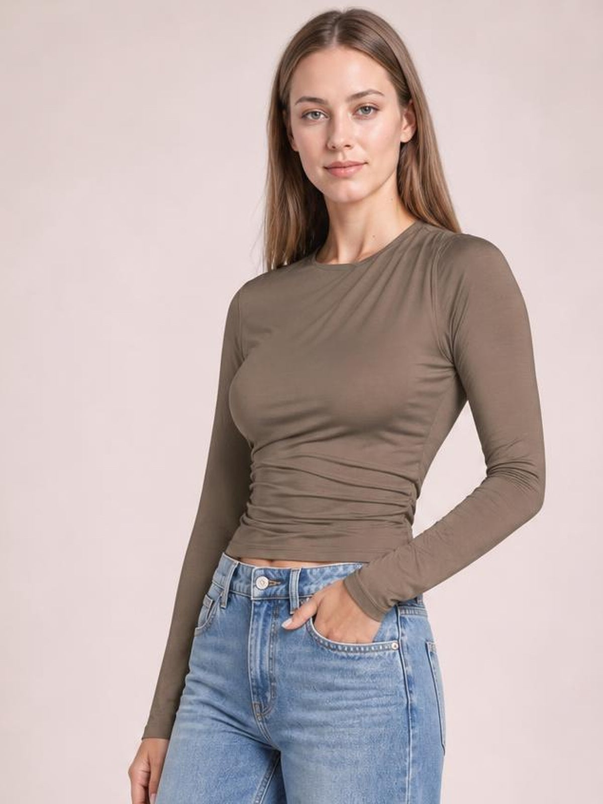 Model wears a taupe ruched asymmetrical-neck long-sleeve T-shirt with a fitted cropped silhouette, gathered waist, and one hand resting in her jeans pocket.