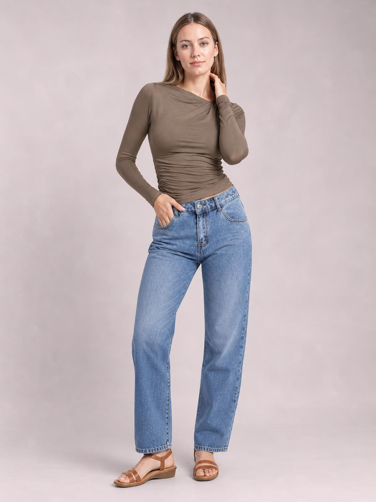 Model wearing a taupe ruched asymmetrical-neck long-sleeve T-shirt with a fitted silhouette, standing with one hand in her jeans pocket.