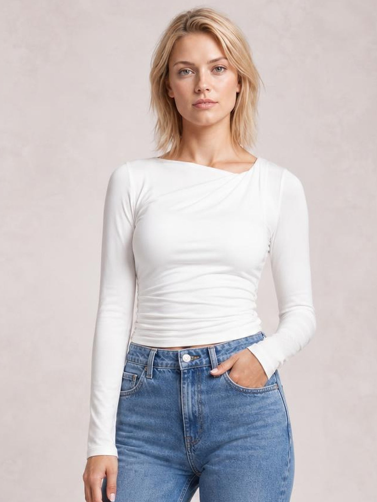 Model wearing a white ruched asymmetrical neck long sleeve T-shirt with a fitted silhouette, paired with blue jeans and one hand in a pocket.