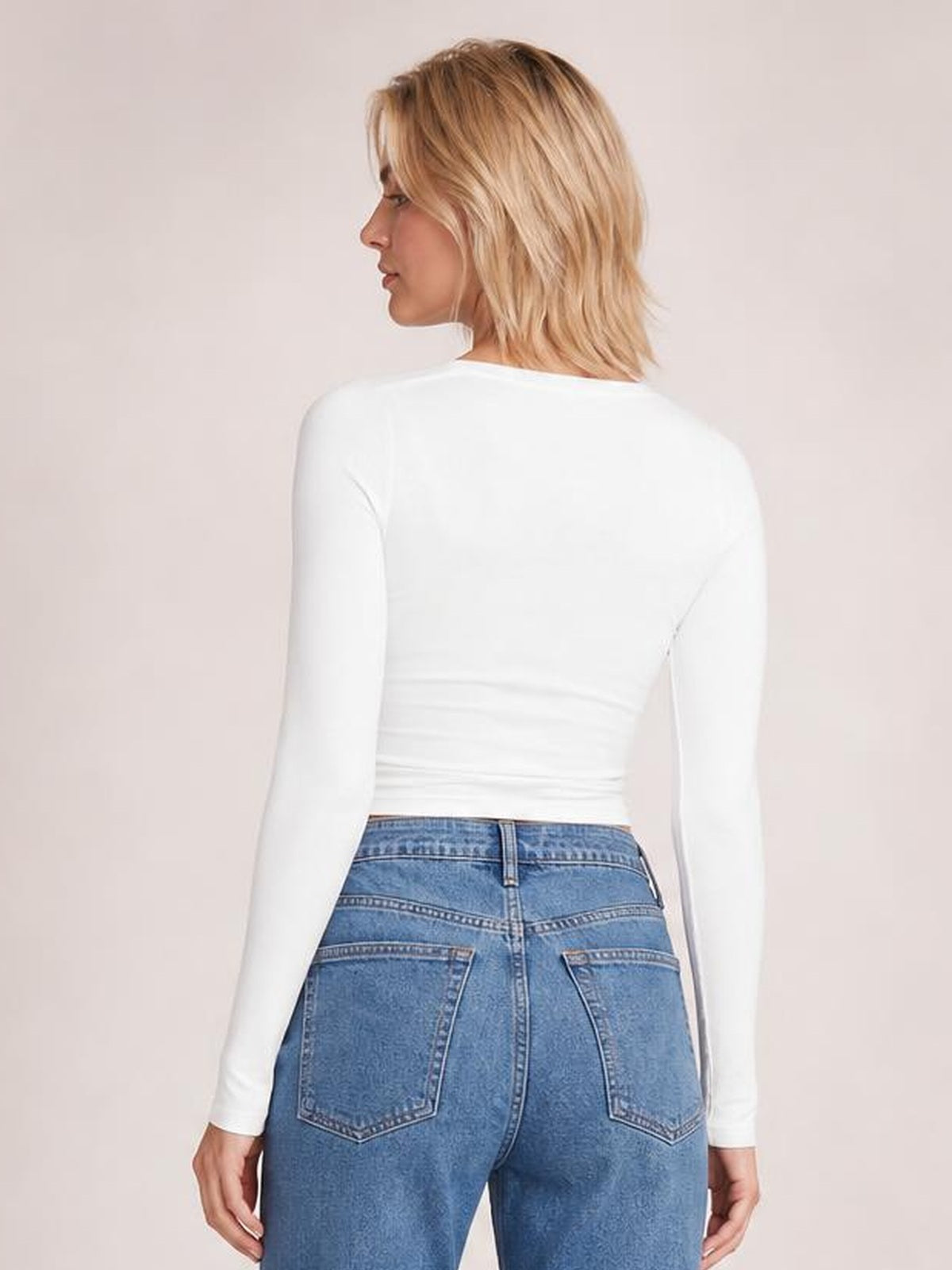 Model in a white long-sleeve T-shirt poses turned away, showing its fitted silhouette and gathered ruching across the lower back.
