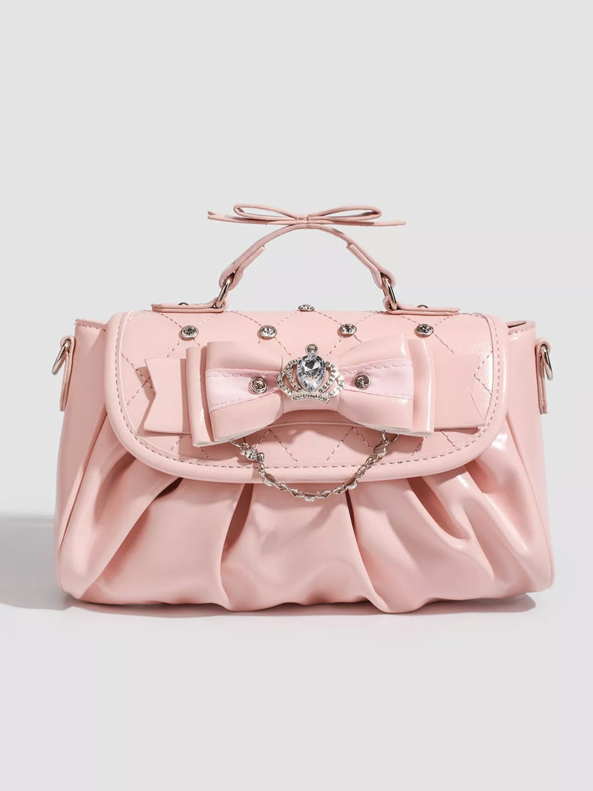 Small pink PU leather crossbody bag with a ruched silhouette, structured top handle, oversized bow, quilted stitching, jeweled clasp, and chain accent.