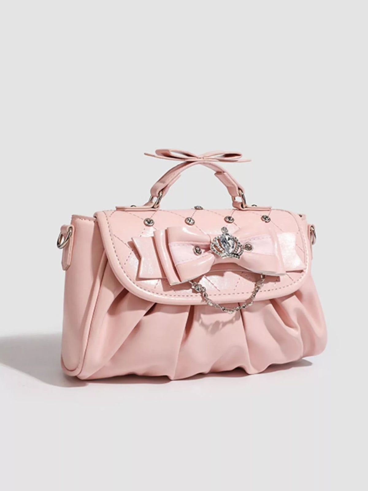 Small pink PU leather crossbody bag with a ruched silhouette, structured flap, oversized bow embellishment, top handles, and decorative chain detail.