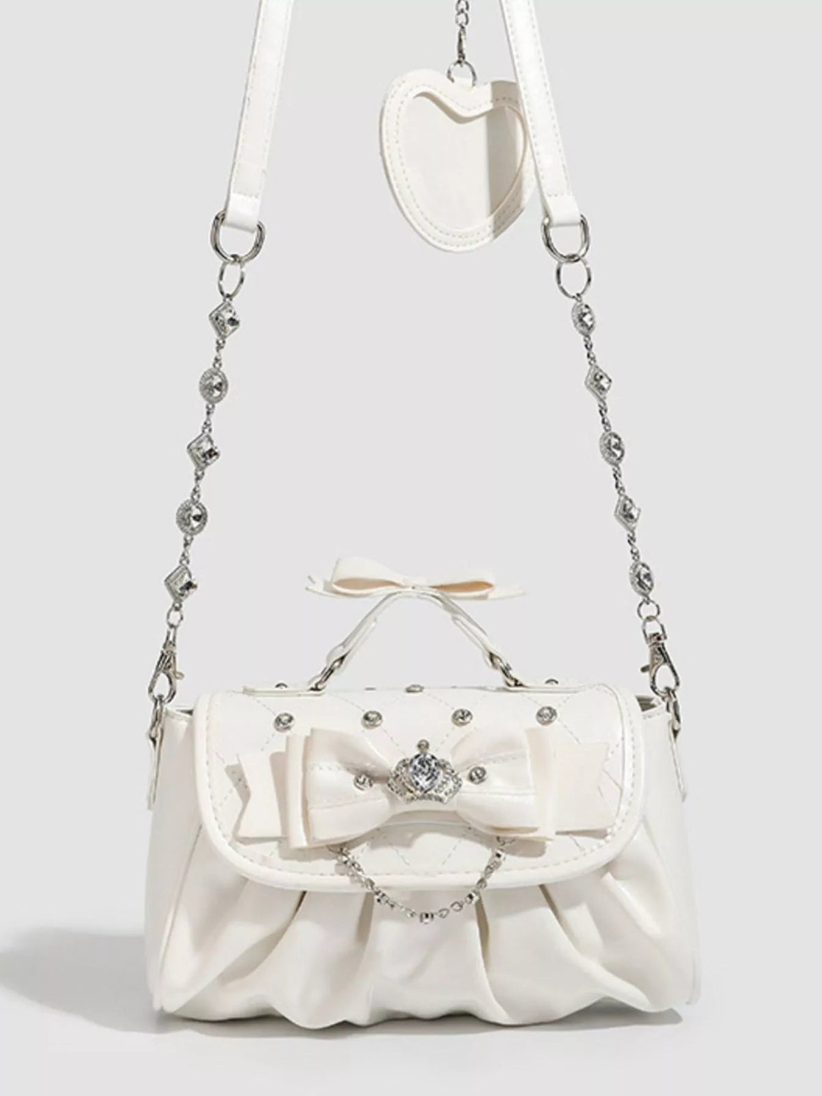 Small white PU leather crossbody bag with a ruched lower body, oversized bow, jeweled embellishment, and decorative chain-and-strap detail.