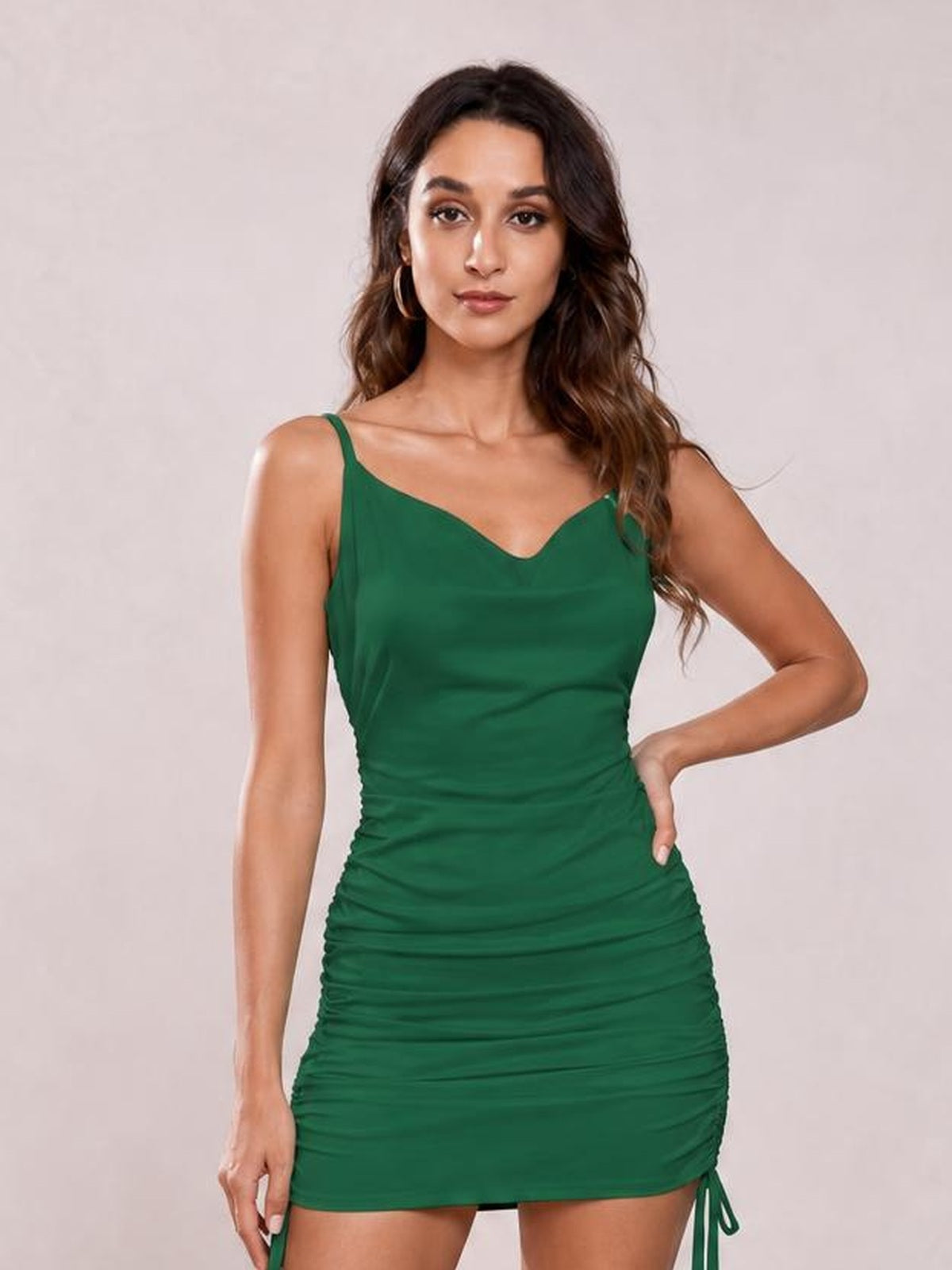A model poses with one hand on her hip in a fitted emerald green ruched cowl neck spaghetti strap mini dress with a gathered silhouette.