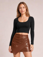 Model stands facing forward in a fitted brown ruched mini skirt with a glossy finish, asymmetrical wrap-style hem, and sleek close-fitting silhouette.