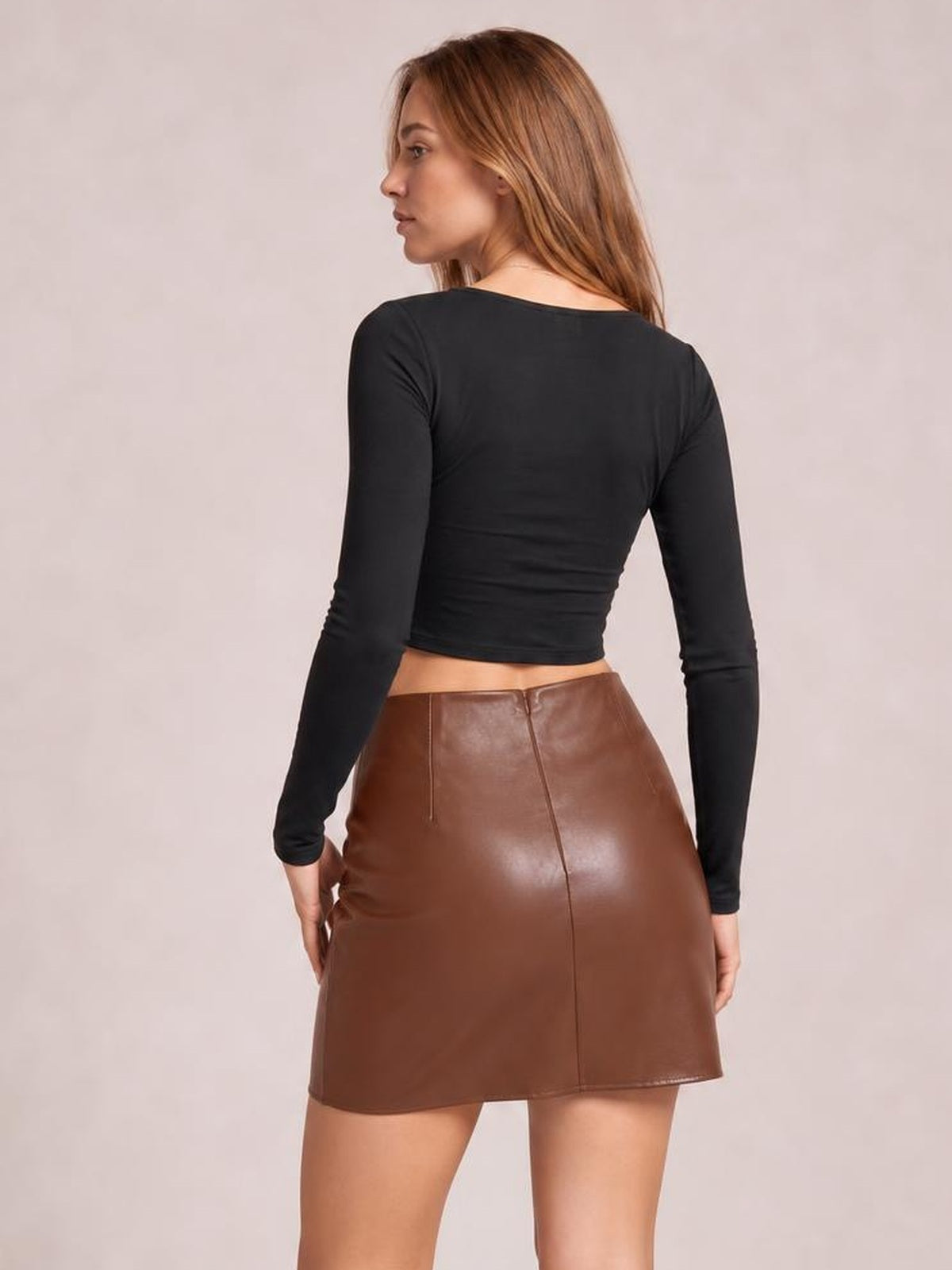 Model shown from behind in a brown fitted mini skirt with a smooth faux-leather look, high waist, center-back seam, and straight silhouette.