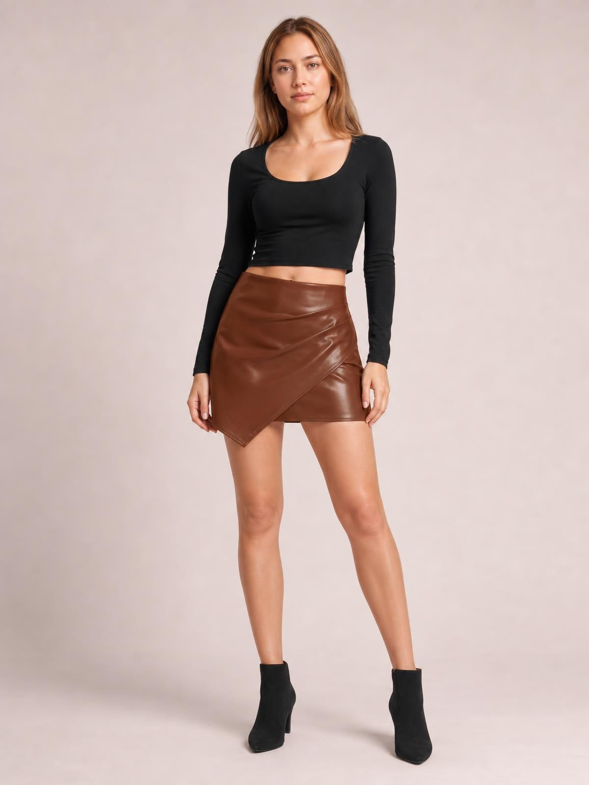 Model stands in a fitted brown ruched mini skirt with a glossy finish, asymmetrical overlapping hem, and a sleek, edgy silhouette.