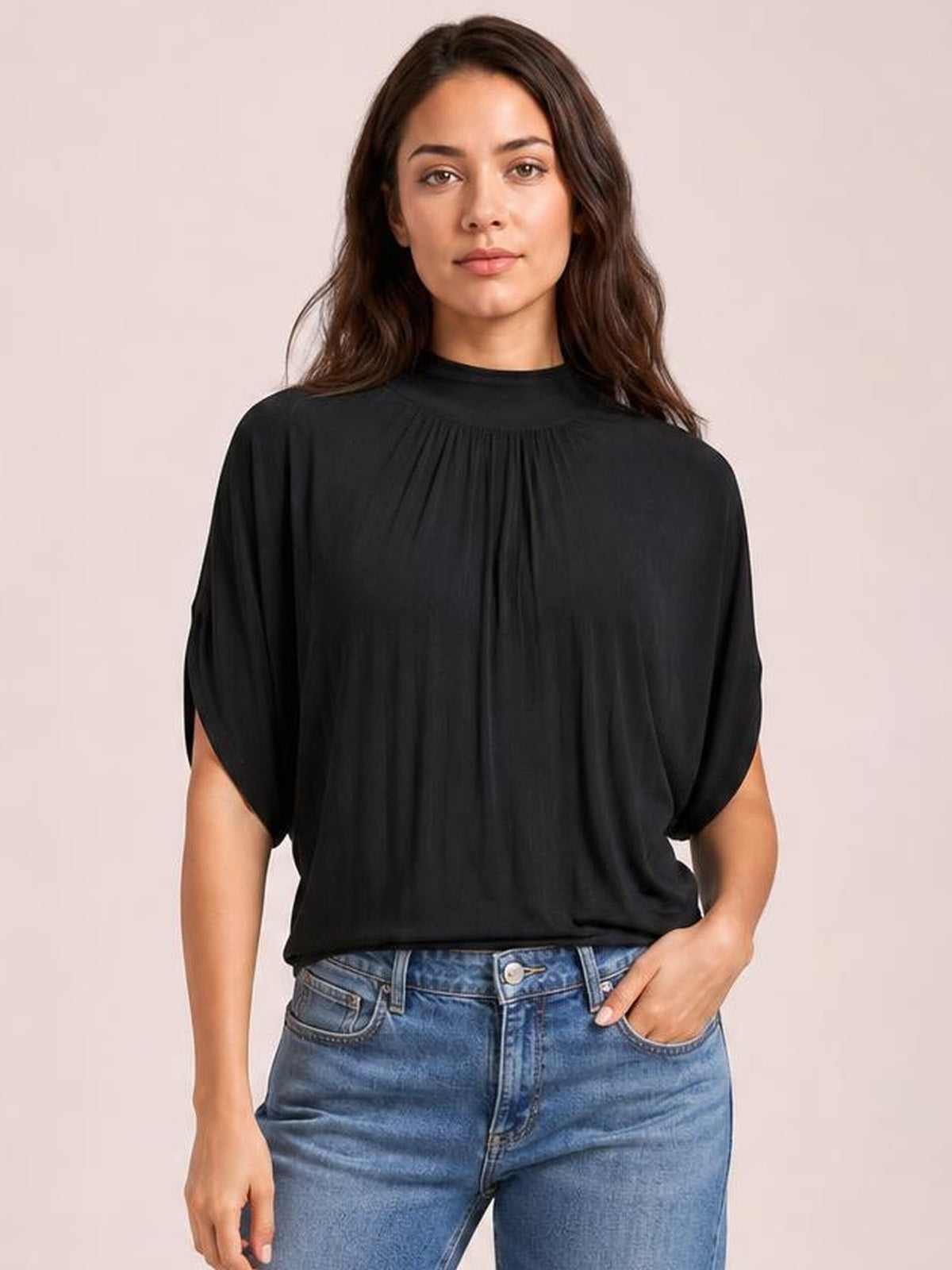 Model wearing a black ruched mock neck blouse with loose half sleeves and a relaxed draped silhouette, standing with one hand in her jeans pocket.