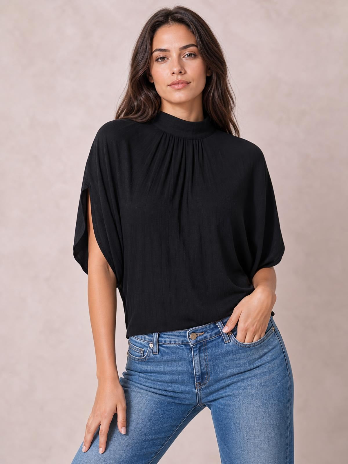 A woman poses in a black ruched mock neck blouse with flowing half sleeves and a relaxed silhouette, paired with blue jeans and one hand in a pocket.