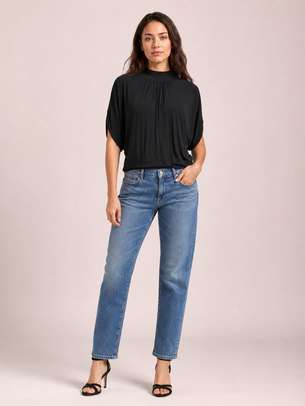 A model stands with one hand in her jeans pocket, wearing a black ruched mock neck blouse with half sleeves and a loose, flowing silhouette.