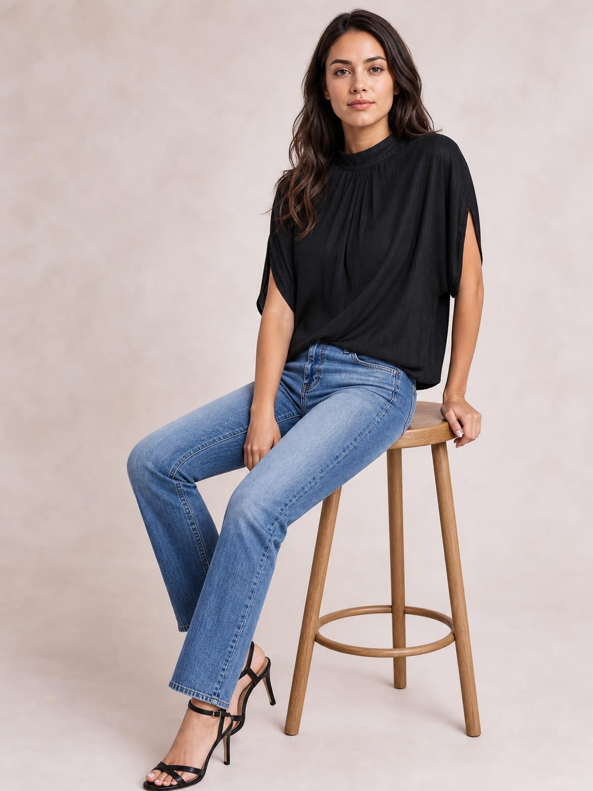 A seated model wears a black ruched mock neck half sleeve blouse with gathered neckline pleats, open sleeve slits, and a loose draped silhouette.