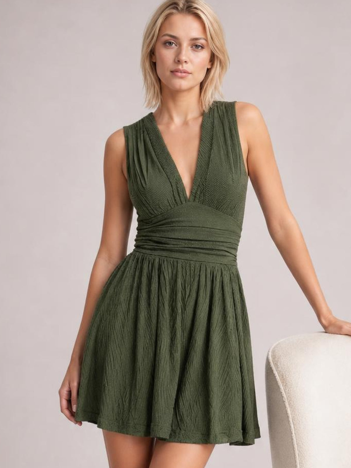 Model poses in an olive green ruched plunge sleeveless romper with a fitted waist, textured construction, and a flared short silhouette.