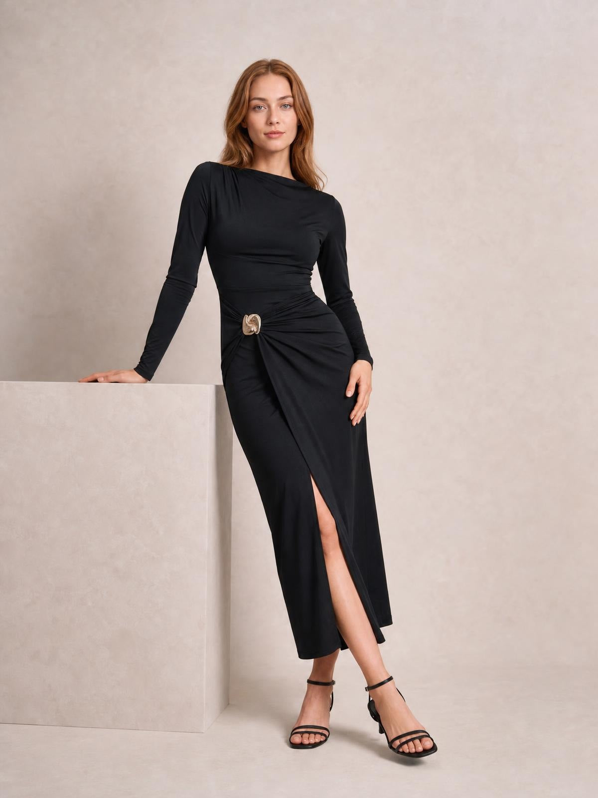 Model poses in a black ruched slit long-sleeve bodycon dress with a decorative waist button and fitted silhouette, resting one hand on a ledge.