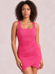 Model poses in a bright pink ruched mini tank dress with a square neckline, wide straps, fitted silhouette, subtle slit, and overlapping asymmetrical hem.