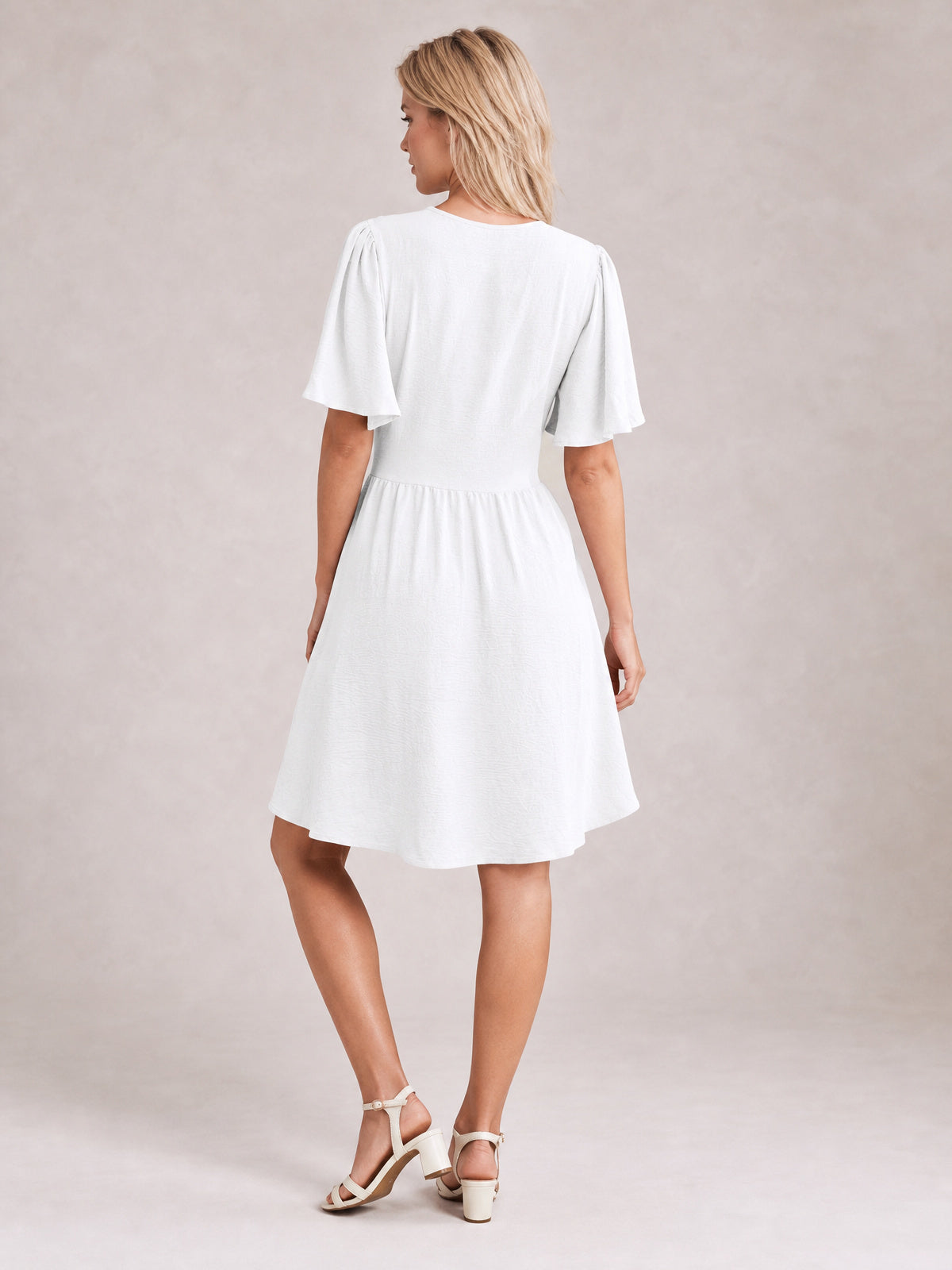 A model wears a white ruched short sleeve dress with fluttered sleeves, a defined waist, and a gathered A-line skirt, posing with one leg crossed.