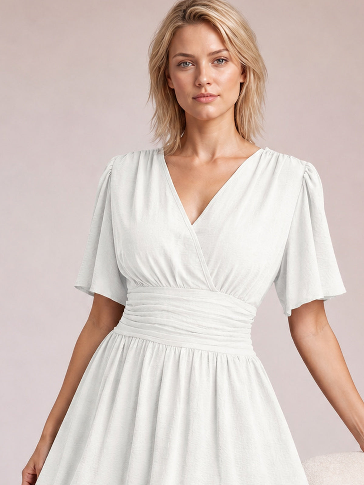 Model wears a white ruched surplice dress with a V-neckline, fluttered short sleeves, defined waistband, and gathered skirt while standing with arms relaxed.