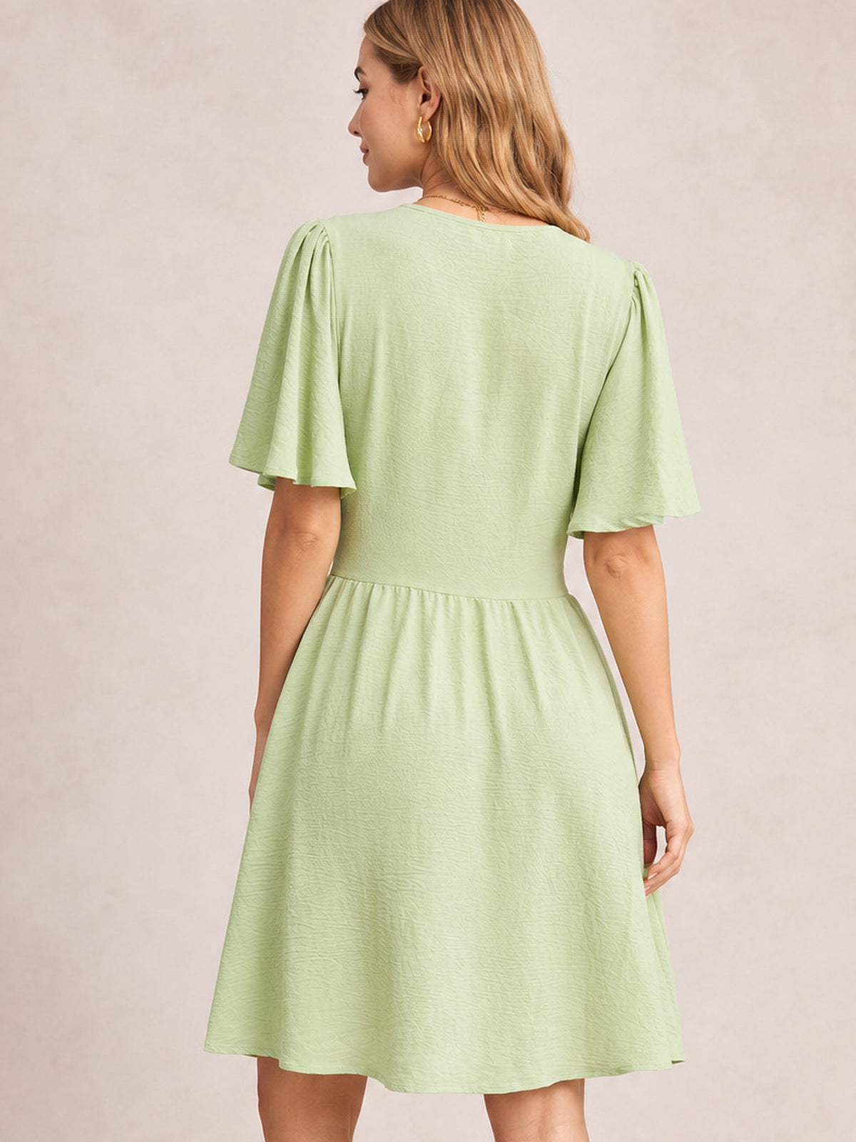 Model turned away in a light green ruched short sleeve dress with fluttered sleeves, a defined waist seam, and a gathered, flared short skirt.