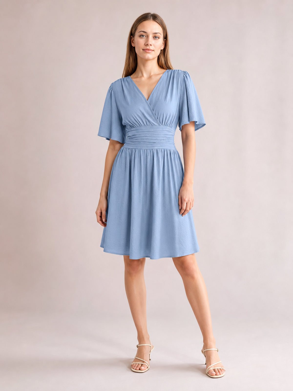 A model poses in a light blue ruched surplice dress with fluttered short sleeves, a defined waistband, and a gathered, flared short skirt.