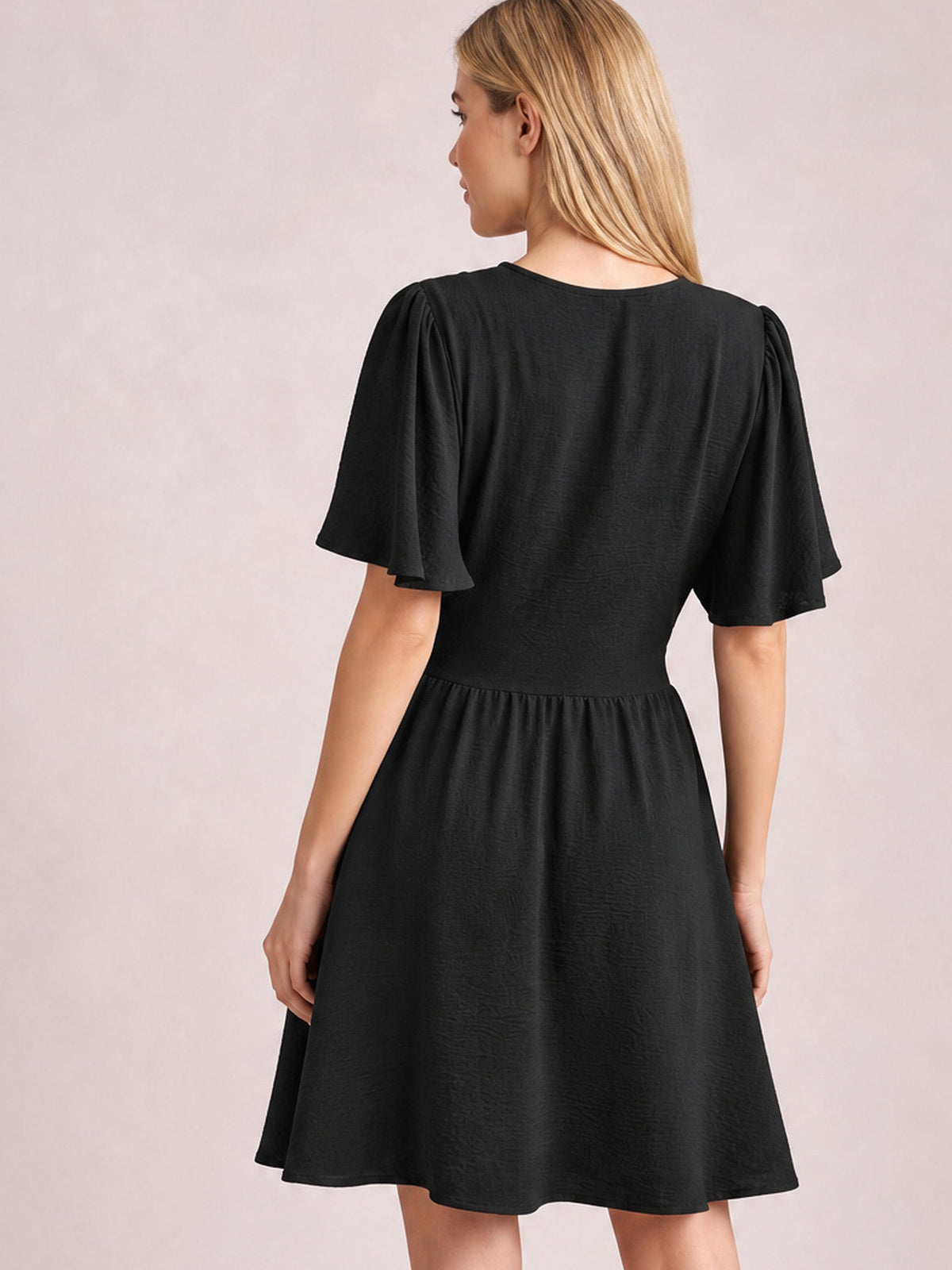 Model shown from the back in a black ruched short-sleeve dress with fluttered sleeves, a defined waistband, and a gathered flared skirt.