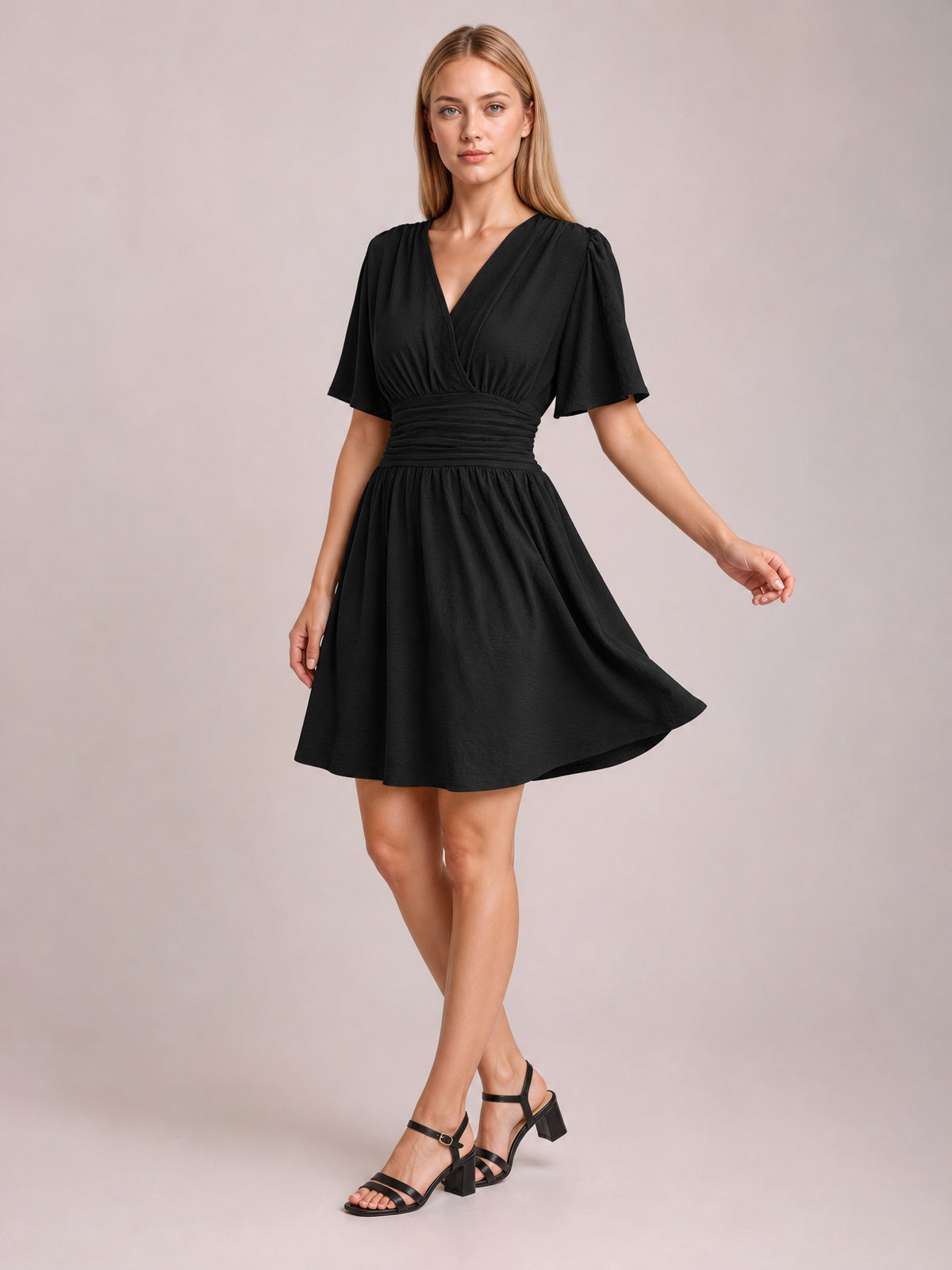 A model poses in a black ruched surplice short sleeve dress with fluttered sleeves, a defined ruched waistband, and a flowing gathered short skirt.
