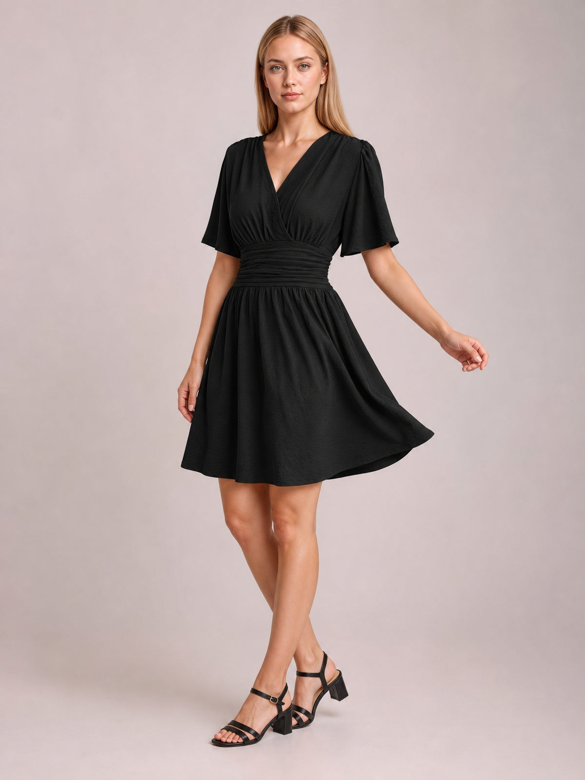 A model poses in a black ruched surplice short sleeve dress with fluttered sleeves, a defined ruched waistband, and a flowing gathered short skirt.