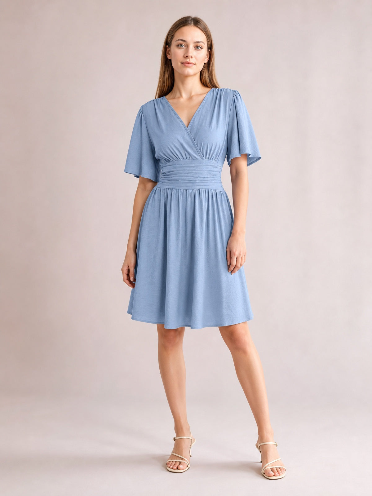 A model poses in a light blue ruched surplice dress with fluttered short sleeves, a defined waistband, and a gathered, flared short skirt.