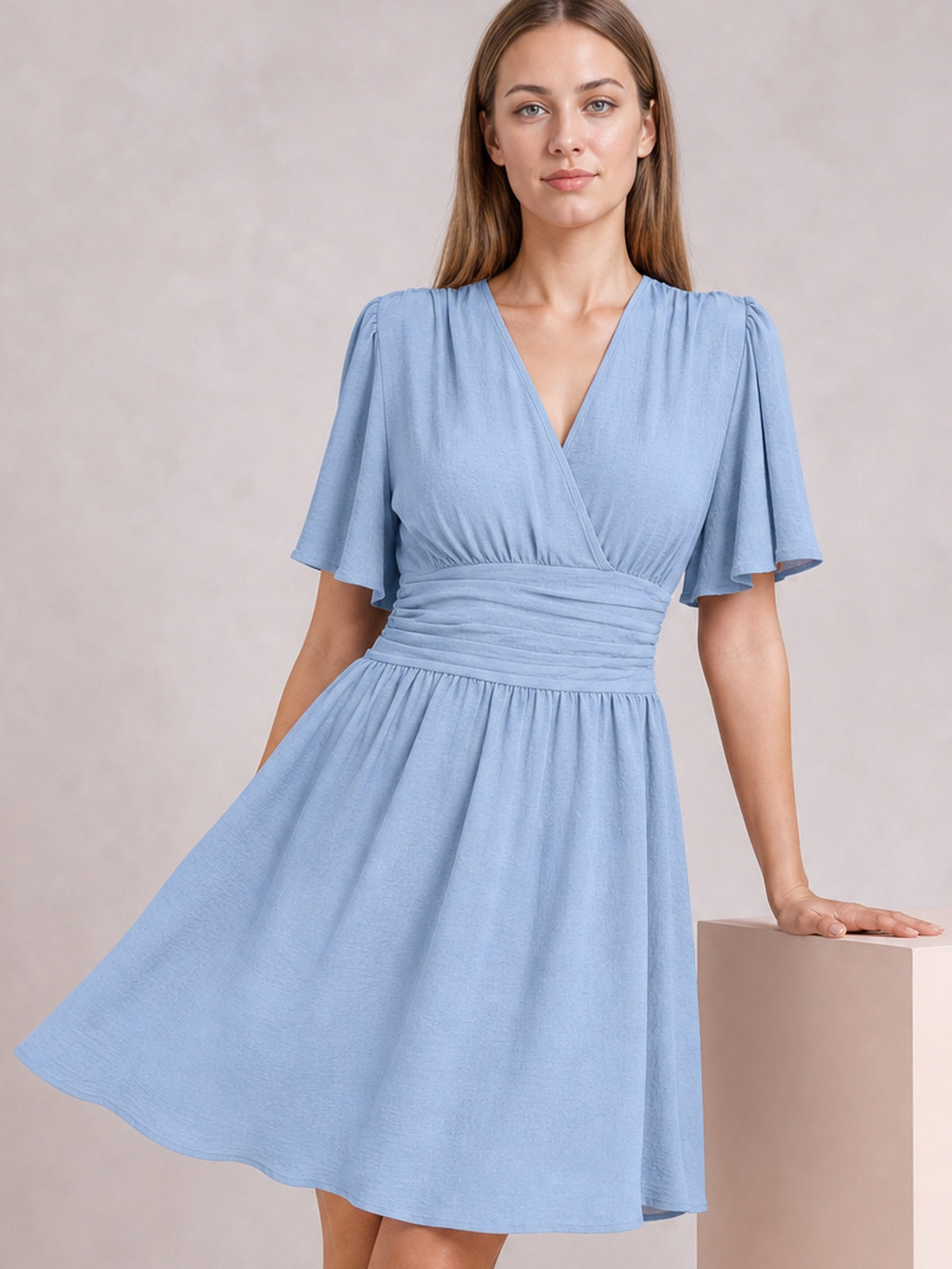 A model poses in a light blue ruched surplice short sleeve dress with fluttered sleeves, a defined waistband, and a flowing gathered skirt.