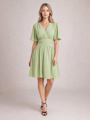 Model wears a light green ruched surplice dress with flutter short sleeves, a defined waistband, and a gathered knee-length skirt, posed with crossed legs.