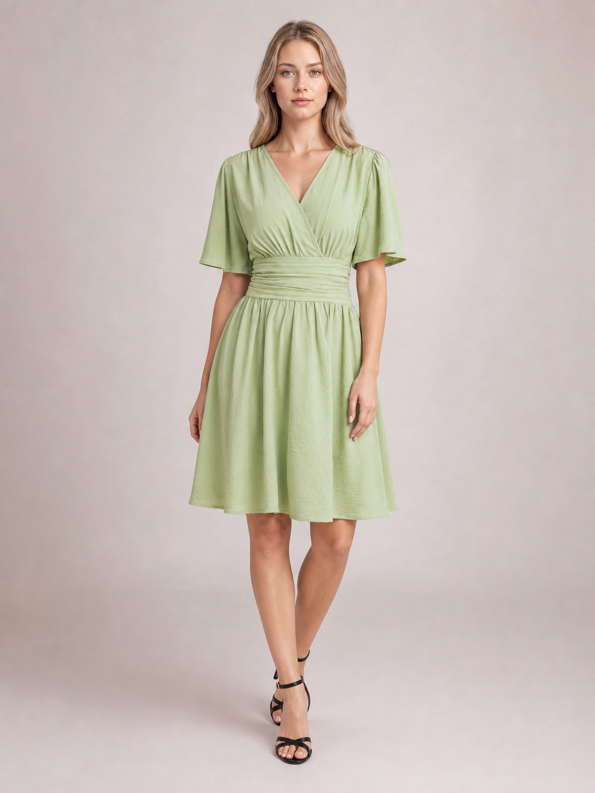 Model wears a light green ruched surplice dress with flutter short sleeves, a defined waistband, and a gathered knee-length skirt, posed with crossed legs.