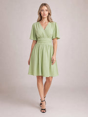 Model wears a light green ruched surplice dress with flutter short sleeves, a defined waistband, and a gathered knee-length skirt, posed with crossed legs.