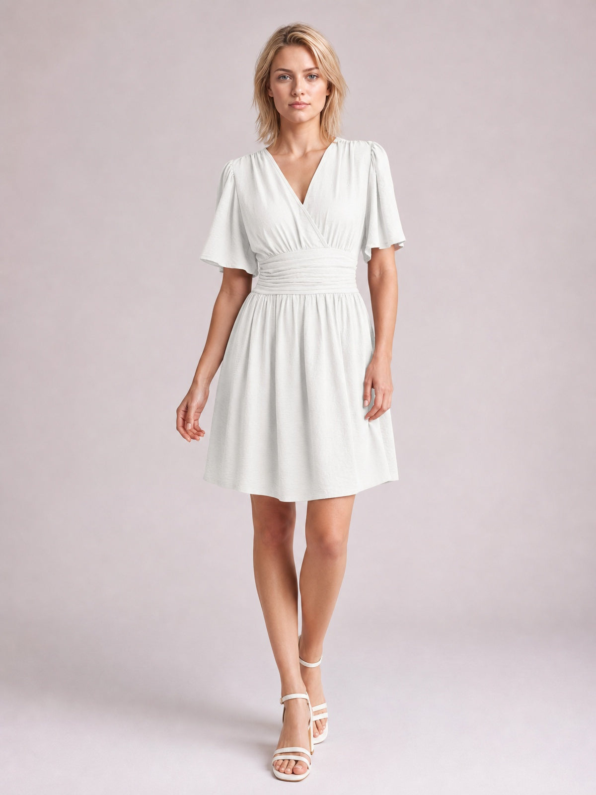 Model wears a white ruched surplice short sleeve dress with fluttered sleeves, defined waistband, gathered A-line skirt, and short hem.