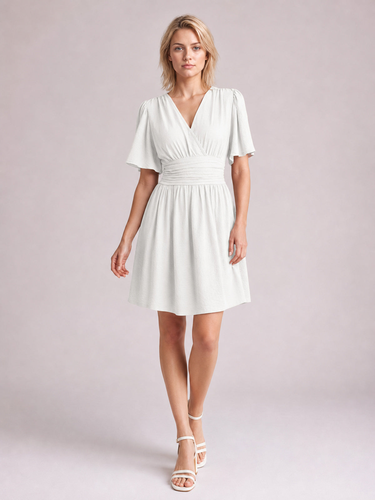 Model wears a white ruched surplice short sleeve dress with fluttered sleeves, defined waistband, gathered A-line skirt, and short hem.